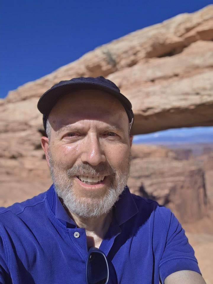 Marshall Gittler, smiling, in a cap and blue shirt in front of a sandstone arch under a deep blue sky